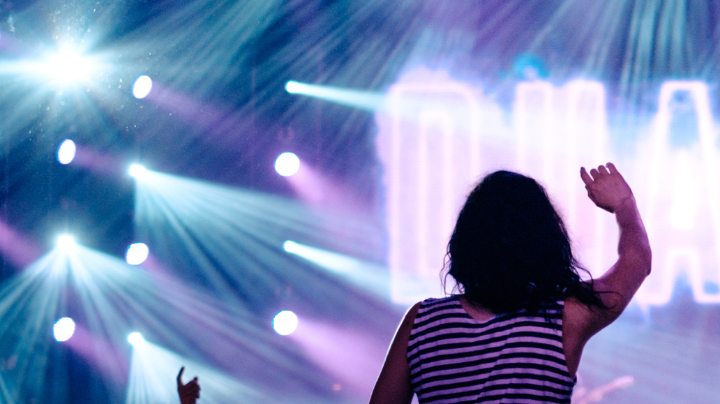 person standing in front of a stage with lights, raising their arms